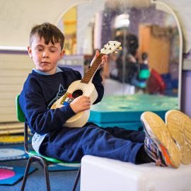 A child seated with their feet up and eyes closed playing a ukulele