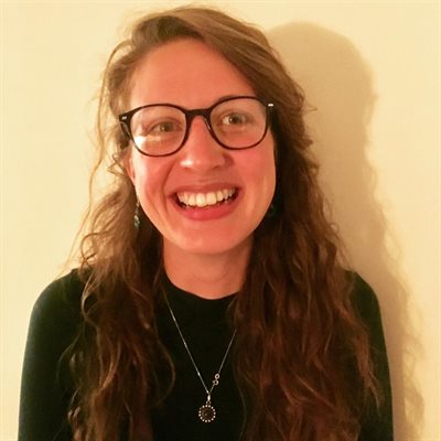 Rosie has long hair, glasses, and is facing the camera, smiling. She is wearing a green top and has long light red hair.