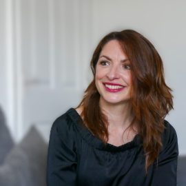 Lady smiling towards stage right of camera with dark hair. She has a book in her hands and is sitting down.