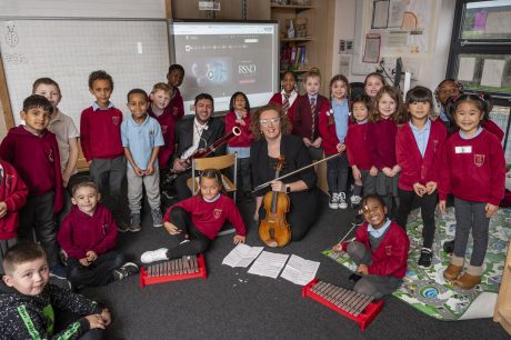 A group of school children smiling at the camera with a woman holding a violin and a man holding a bassoon