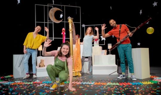 Four people smiling and waving surrounded by a variety of instruments and nighttime images.