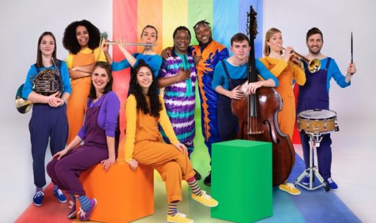 10 people wearing colourful clothes holding a variety of instruments in front of a rainbow background.