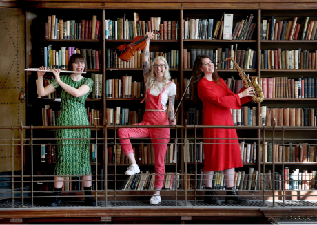 Three musicians wearing bright clothes stand in front of library bookshelves holding a flute, violin and saxophone.